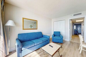 A living room with a blue sofa, a smaller blue armchair, a coffee table, and a lamp. - Camelot by the Sea 1704 (Myrtle Beach)