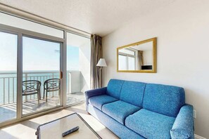 A room with a blue sofa, a glass door leading to a balcony with chairs, and a mirror on the wall. - Camelot by the Sea 1704 (Myrtle Beach)