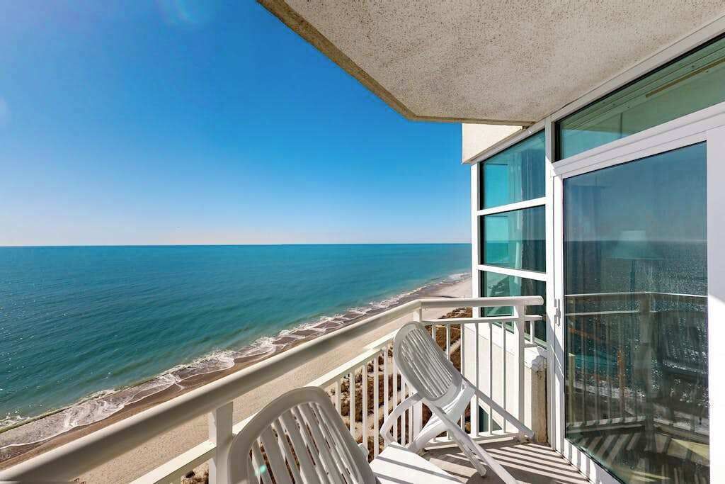 A balcony with a view of the ocean, white chairs, and a glass door.