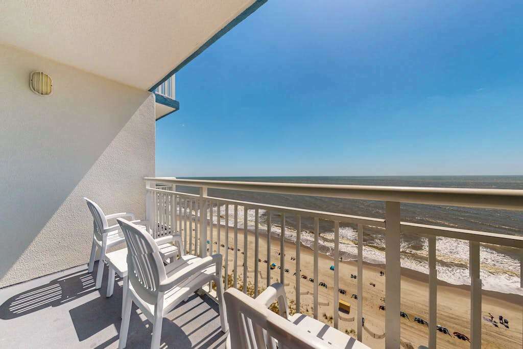 A balcony with a view of the beach, white chairs, and a railing.