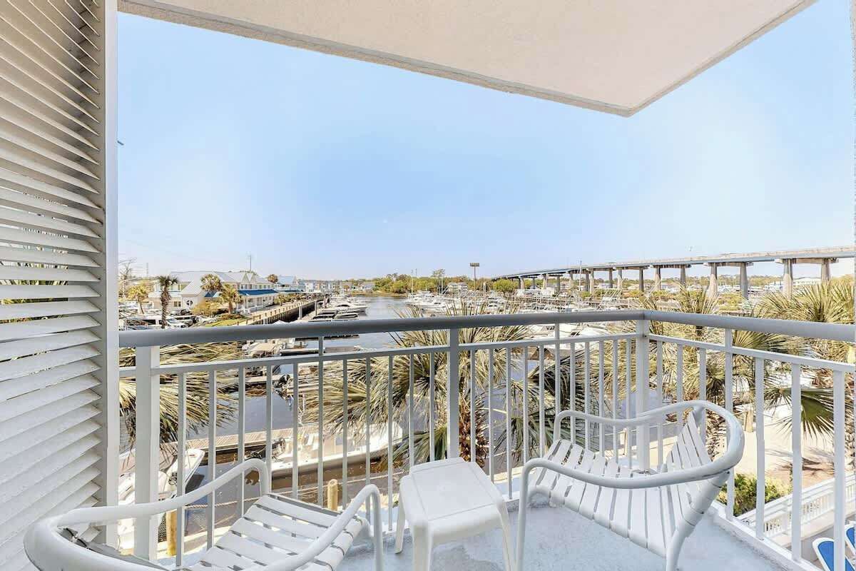 A balcony with white railings and chairs, overlooking a marina with boats and palm trees.