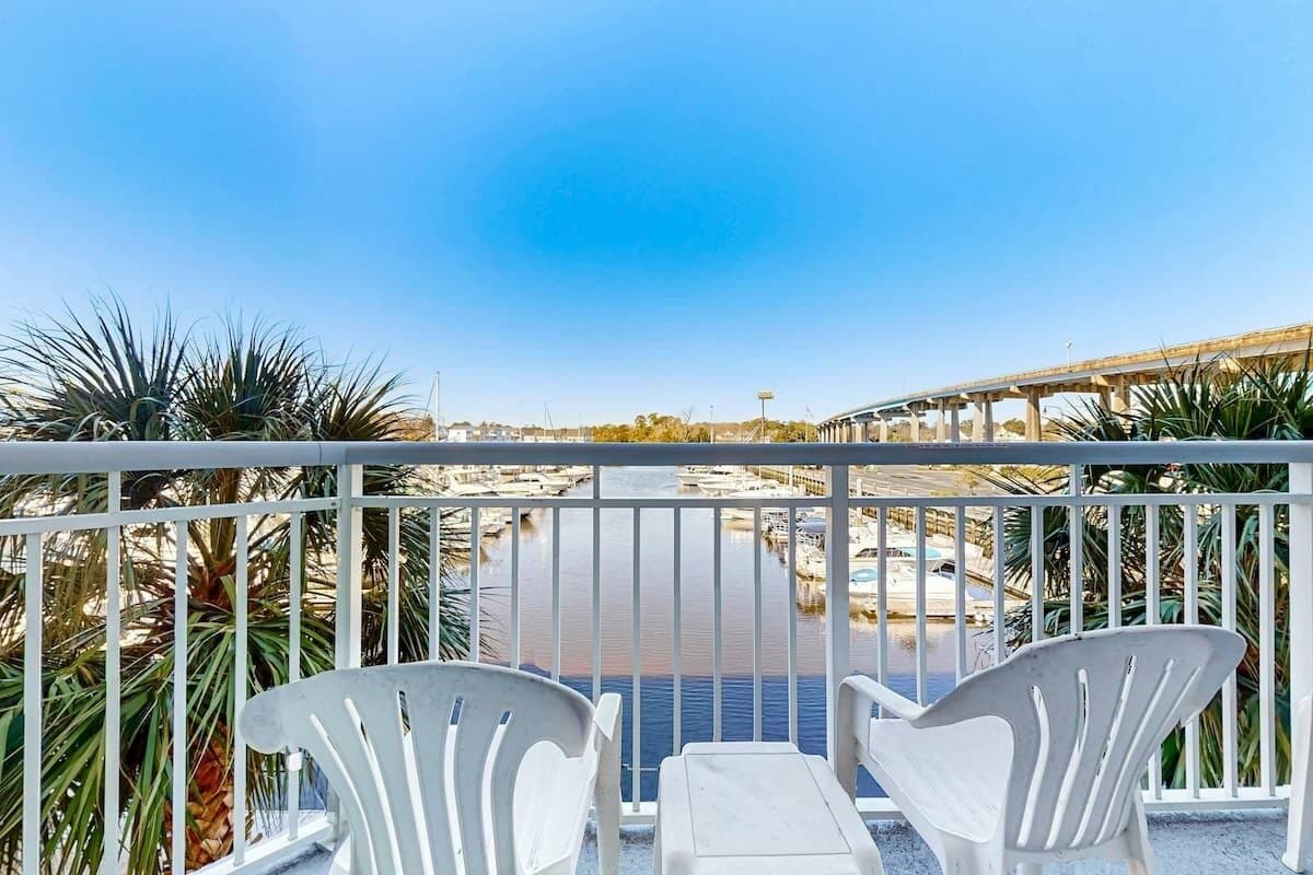 A balcony with white chairs and a table, overlooking a marina with boats and a clear blue sky.