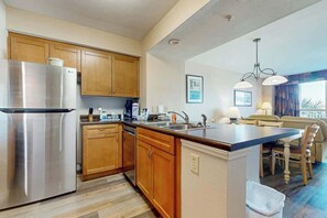 A modern kitchen with wooden cabinets, a stainless steel refrigerator, and a dining area with a table and chairs. - Harbourgate Unit 0130 (North Myrtle Beach)