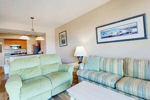 A living room with a sofa, a coffee table, and a lamp. There is a kitchen area in the background with cabinets and appliances. - Harbourgate Unit 0130 (North Myrtle Beach)