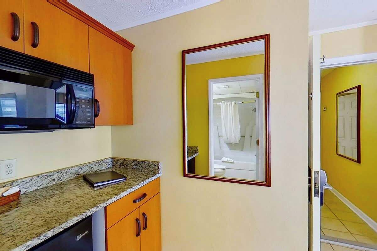 A kitchen with a microwave, granite countertop, wooden cabinets, and a mirror reflecting a bathroom.