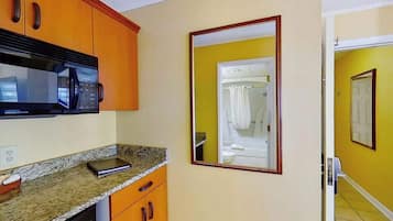 A kitchen with a microwave, granite countertop, wooden cabinets, and a mirror reflecting a bathroom.
