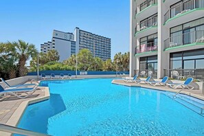 Ein Pool mit Liegestühlen, ein Gebäude mit Balkonen und Palmen.