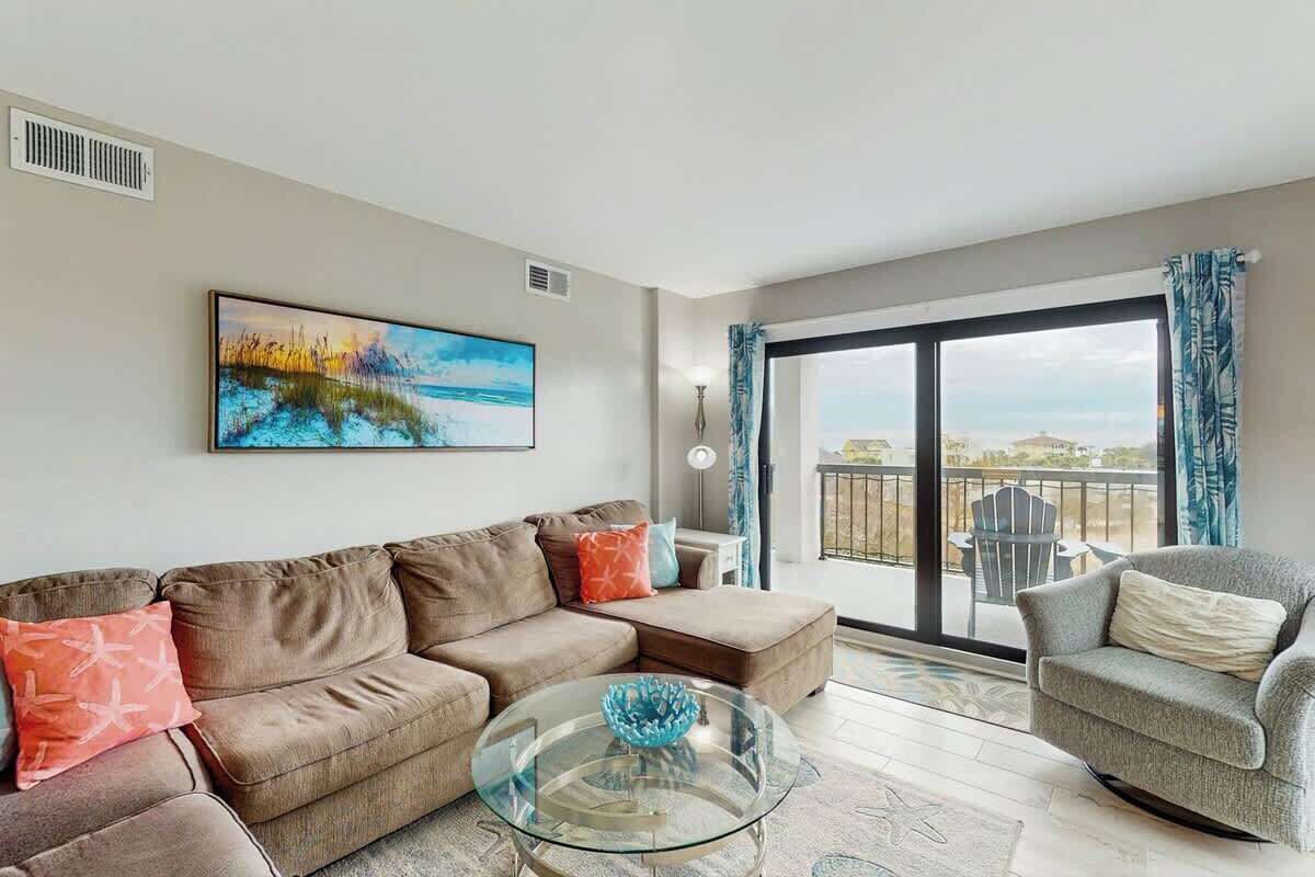 A living room with a sofa, armchair, and glass coffee table. There is a painting on the wall and a view through a large window.