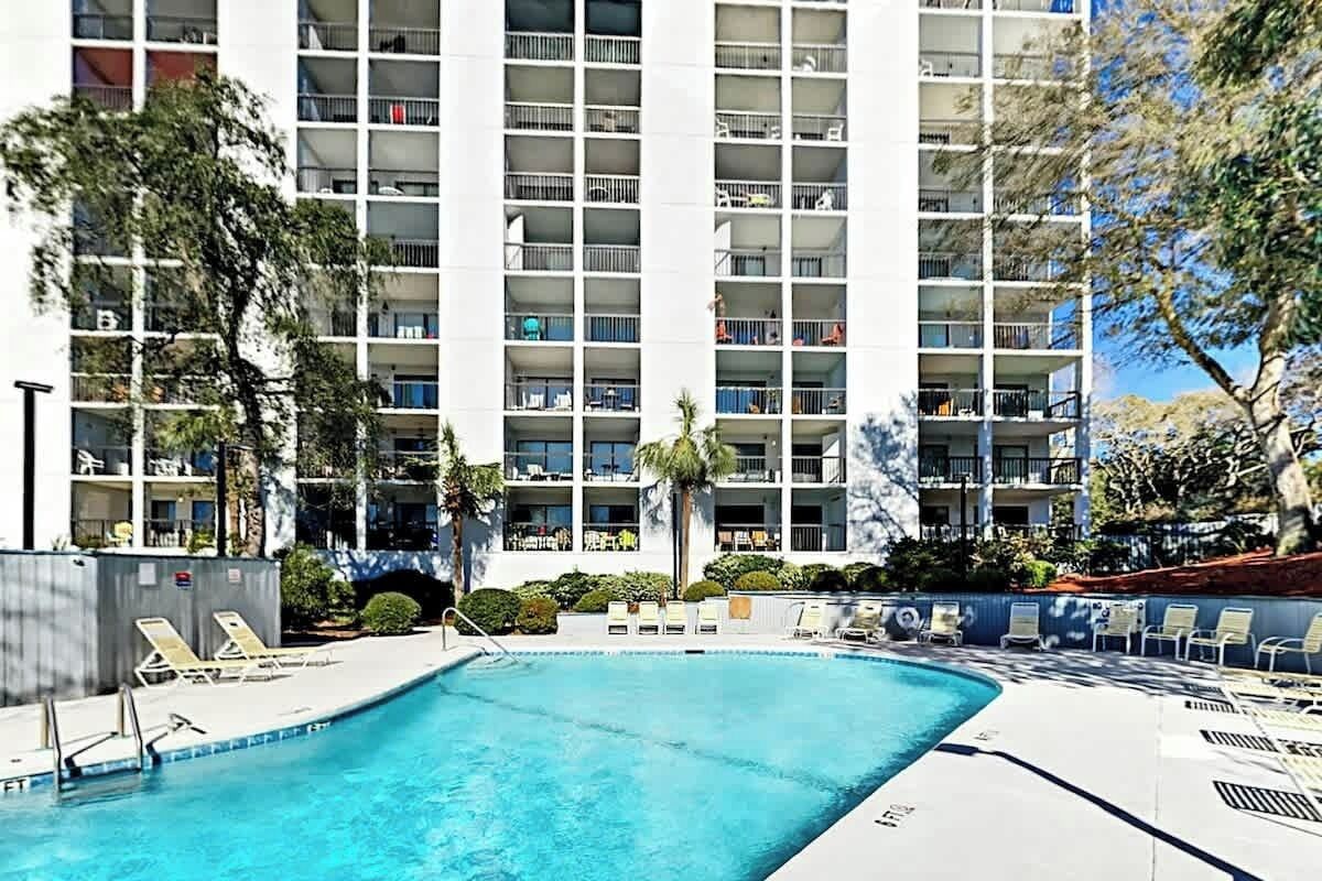 A swimming pool with lounge chairs and a multi-story building with balconies in the background.