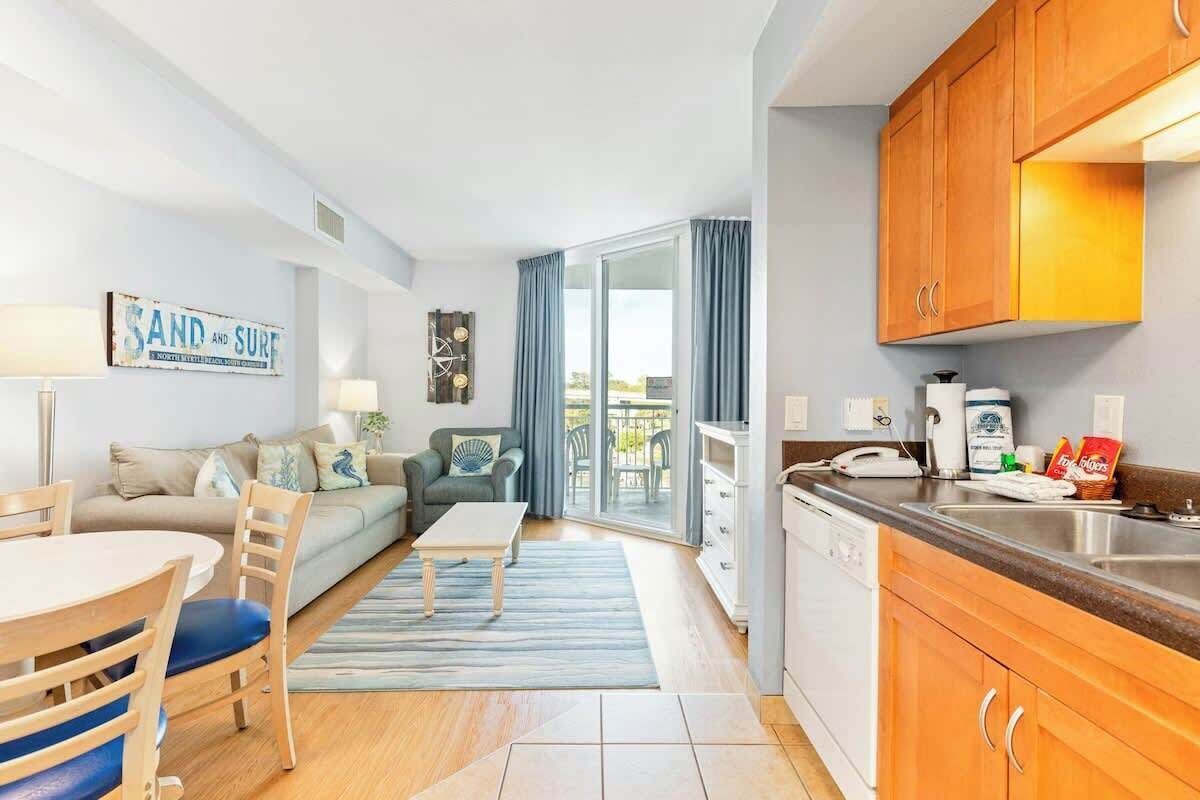 A modern living room with a sofa, coffee table, and chairs, a kitchen with wooden cabinets and a sink, and a balcony with blue curtains.
