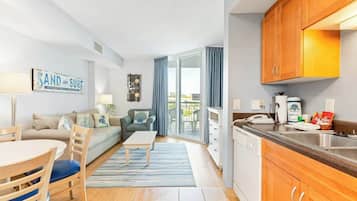 A modern living room with a sofa, coffee table, and chairs, a kitchen with wooden cabinets and a sink, and a balcony with blue curtains.