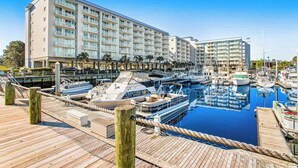 Uma marina com vários barcos atracados, um prédio de hotel grande com varandas, e um céu azul e límpido.