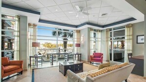 A spacious living area with a sofa, armchairs, a coffee table, and a TV. There are large windows offering a view of palm trees and a balcony. - Harbourgate Unit 0522 (North Myrtle Beach)