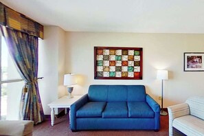 A room with a blue sofa, a white table with a lamp, a patterned artwork on the wall, and a window with curtains. - Harbourgate Unit 0626 (North Myrtle Beach)