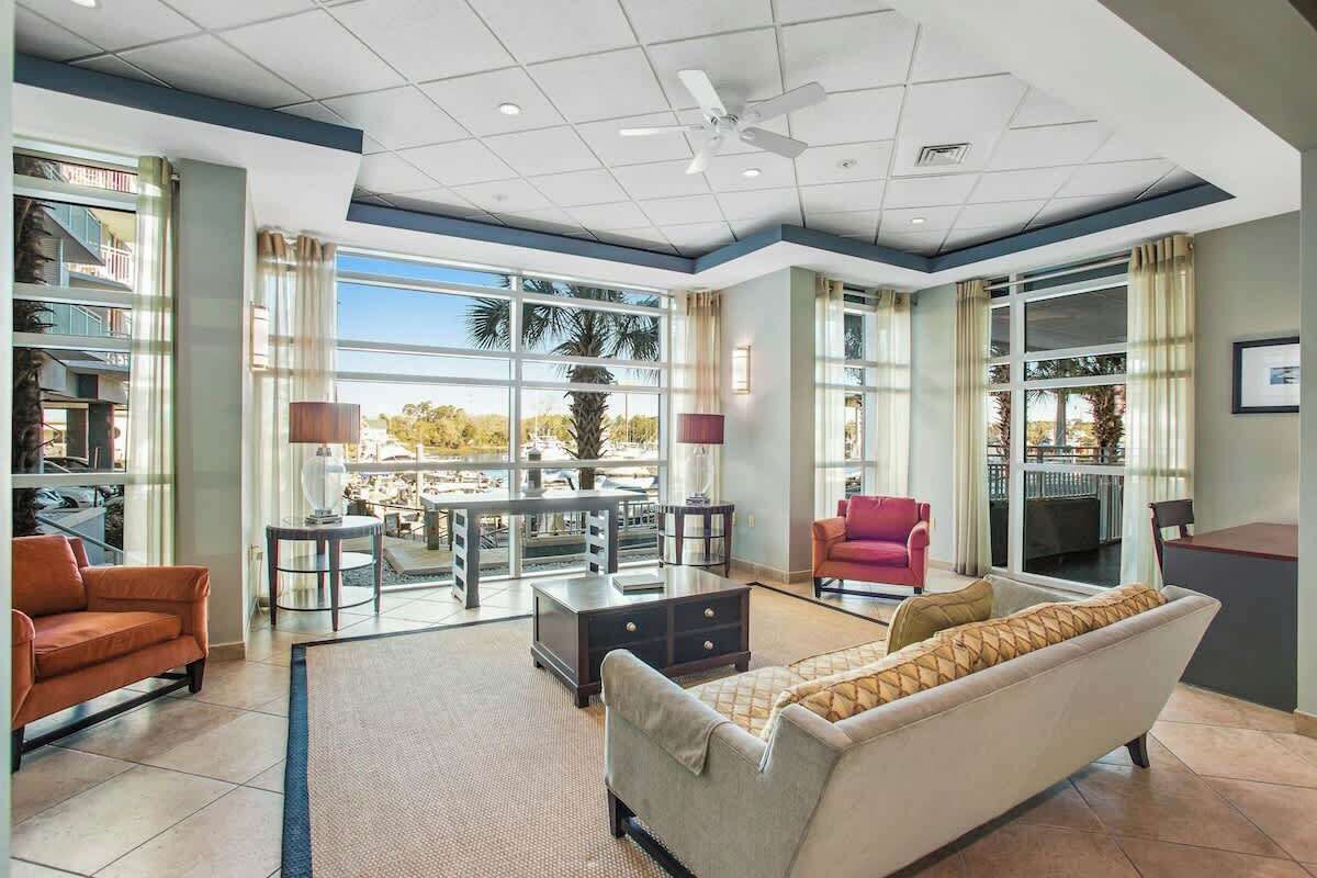 Un vaste espace de vie comprenant un canapé, des fauteuils, une table basse et un téléviseur. De grandes fenêtres offrent une vue sur les palmiers, et un balcon est également disponible.