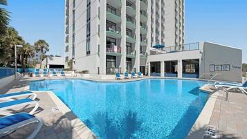 Uma área de piscina de hotel com uma piscina azul cristalina, espreguiçadeiras e um edifício alto com varandas ao fundo.