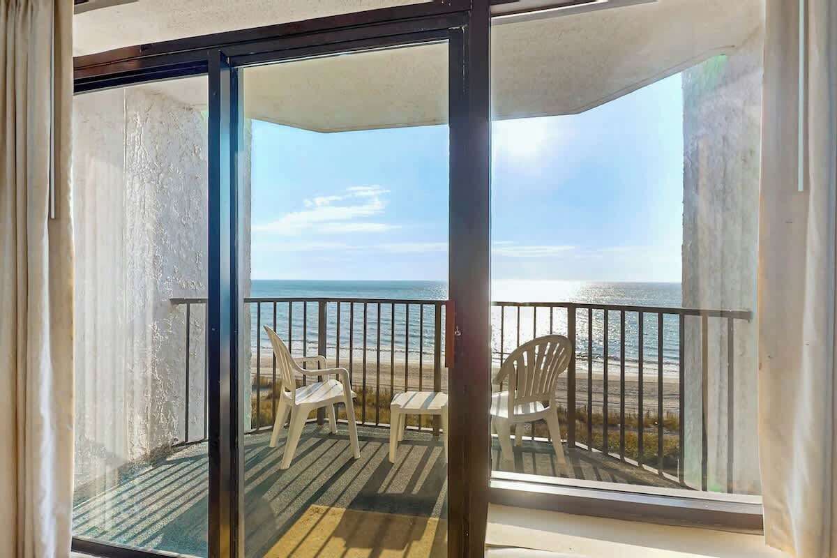 A balcony with a view of the beach, featuring a glass door, a wooden deck, and two white chairs.