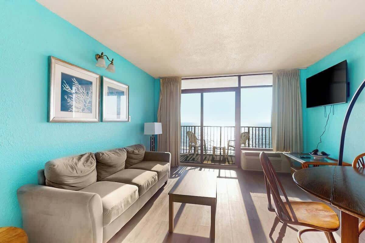 Una habitación con sofá, mesa de comedor, sillas, televisor y balcón con vistas.