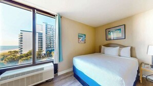 A hotel room with a large bed, a view of the cityscape, and a window with curtains. - Ocean Park Resort 0522 (Myrtle Beach)
