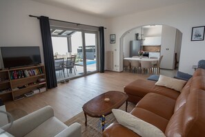 Un salon avec un canapé en cuir, une table basse et une bibliothèque. Une porte-fenêtre coulissante donne accès à un espace extérieur avec une piscine.