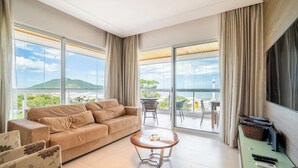 A spacious living room with a beige sofa, a round coffee table, and a balcony with a view of greenery and buildings.