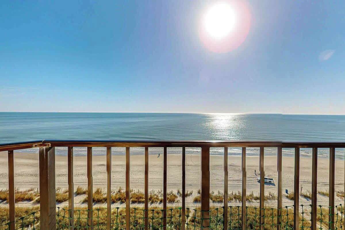 A beach view from a balcony with a railing, a clear sky, and the sun reflecting on the water.