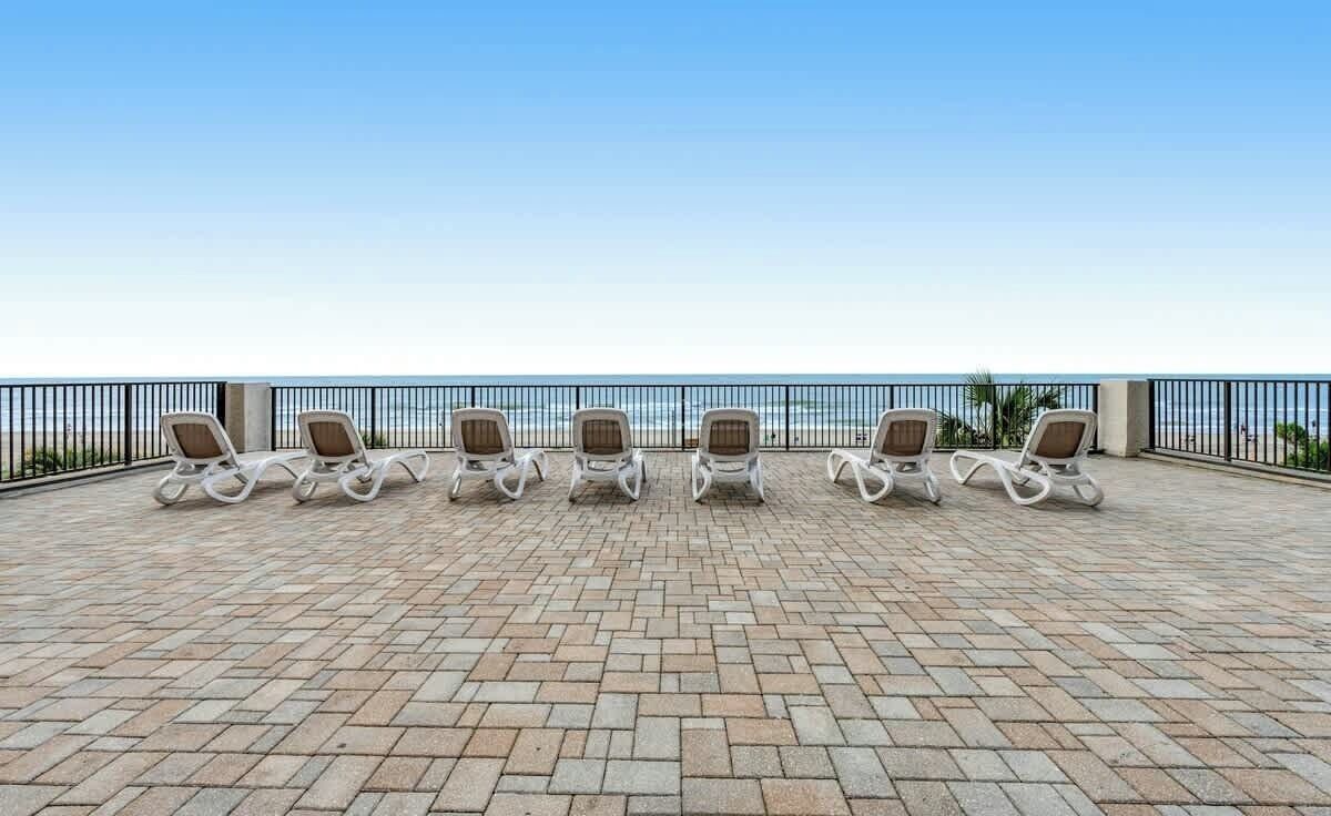 A terrace with six lounge chairs aligned in a row, overlooking a beach.