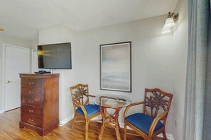 A room with a wooden dresser, a flat-screen TV mounted on the wall, two wicker chairs with blue cushions, and a round glass table. - Ocean Park Resort 1224 (Myrtle Beach)