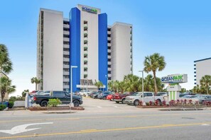 A multi-story hotel with a prominent blue and white facade, a parking lot with various vehicles, and palm trees in the vicinity. - Ocean Park Resort 1224 (Myrtle Beach)