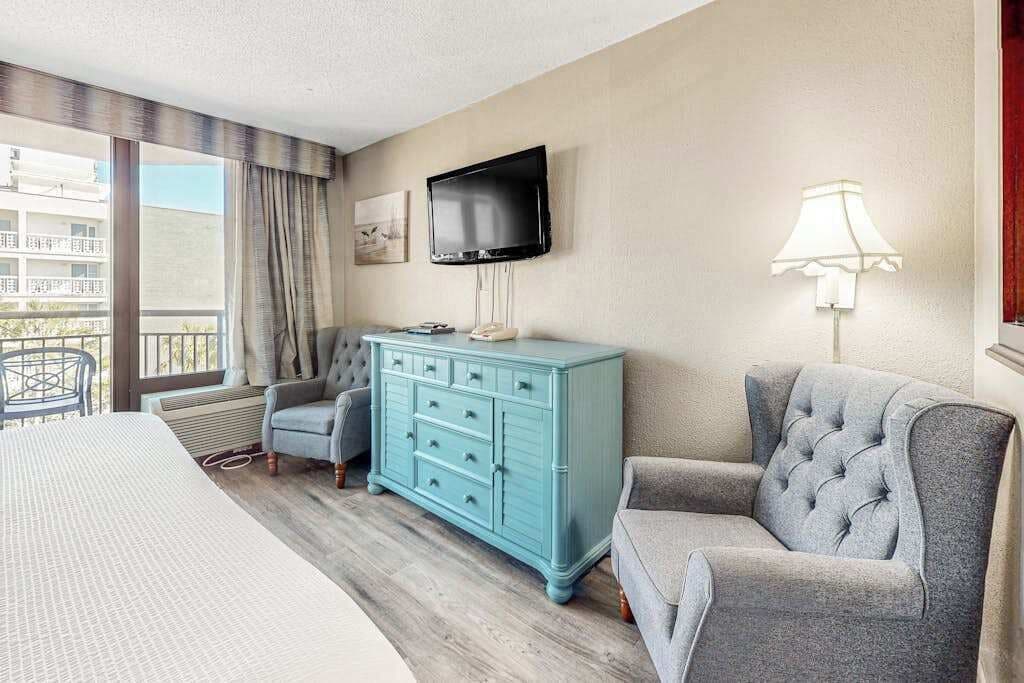 A hotel room with a bed, a blue dresser, a grey armchair, a television, and a window with curtains.