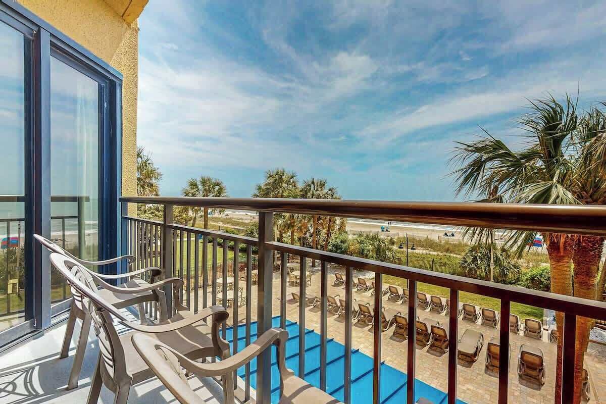 A balcony with a pool, lounge chairs, and a view of palm trees and a distant landscape.