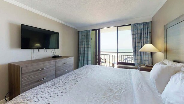 A hotel room with a bed, a TV mounted on the wall, a desk with a lamp, and a view of the outside through a window with curtains. - The Patricia Grand 0624 (Myrtle Beach)