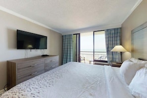 A hotel room with a bed, a TV mounted on the wall, a desk with a lamp, and a view of the outside through a window with curtains. - The Patricia Grand 0624 (Myrtle Beach)