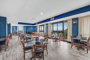 A spacious dining area with multiple tables and chairs, large windows offering a view, and a blue and white color scheme. - The Patricia Grand 1238 (Myrtle Beach)