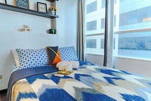 A hotel room with a large bed, a window with blinds, a shelf with decorative items, and a wall with a framed quote. - Comfortable City Escape (Taguig City)