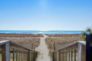 Ein Holzsteg führt zu einem Strand unter wolkenlosem Himmel.