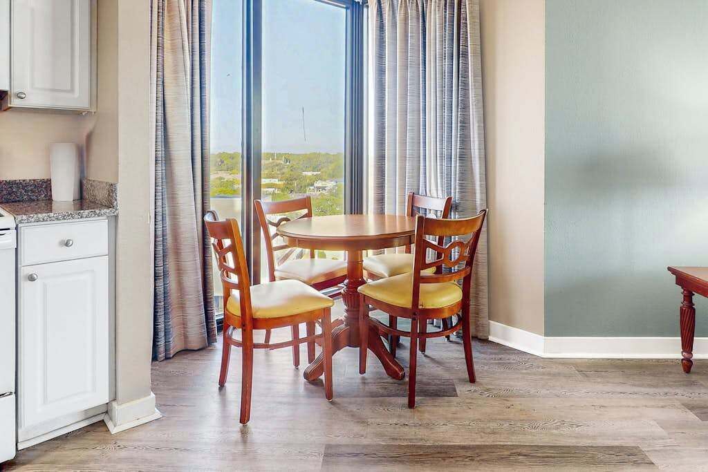 A dining area with a round table and four chairs, a kitchen with white cabinets, and a view through large windows.