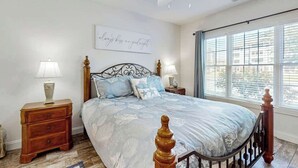 A bedroom with a bed, two bedside tables, a wall-mounted light fixture, and a window with blinds. - Magnolia Pointe Unit 103 (Myrtle Beach)