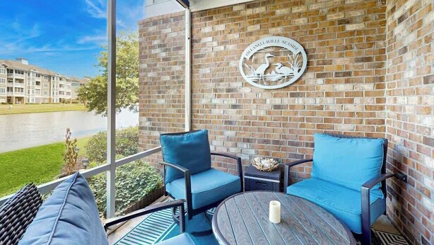 A balcony with a view of a building and a grassy area. There is a round table with a candle and two chairs with blue cushions. - Magnolia Pointe Unit 103 (Myrtle Beach)