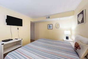 A hotel room with a bed, a flat-screen TV mounted on the wall, a bedside table with a lamp, and a framed picture on the wall. - Waipani Unit C-15 Condo (North Myrtle Beach)
