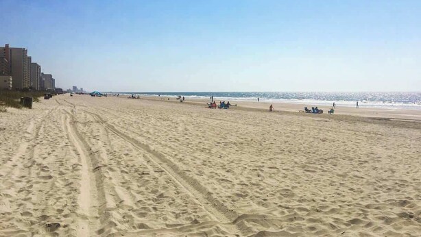 Ein weiter Sandstrand mit Spuren von Autoreifen, einem klaren Himmel und Gebäuden in der Ferne.