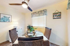 A dining area with a round table, wicker chairs, a ceiling fan, and a window with blinds. - Waipani Unit C-15 Condo (North Myrtle Beach)