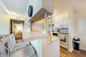 A compact kitchen with white cabinetry, a microwave, an oven, and a small dining area with a white table and chairs. - Waipani Unit C-15 Condo (North Myrtle Beach)