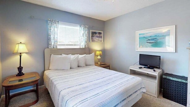 A bedroom with a bed, bedside tables, a television, and a window with curtains. - Dunes Pointe Unit C-14 (Myrtle Beach)