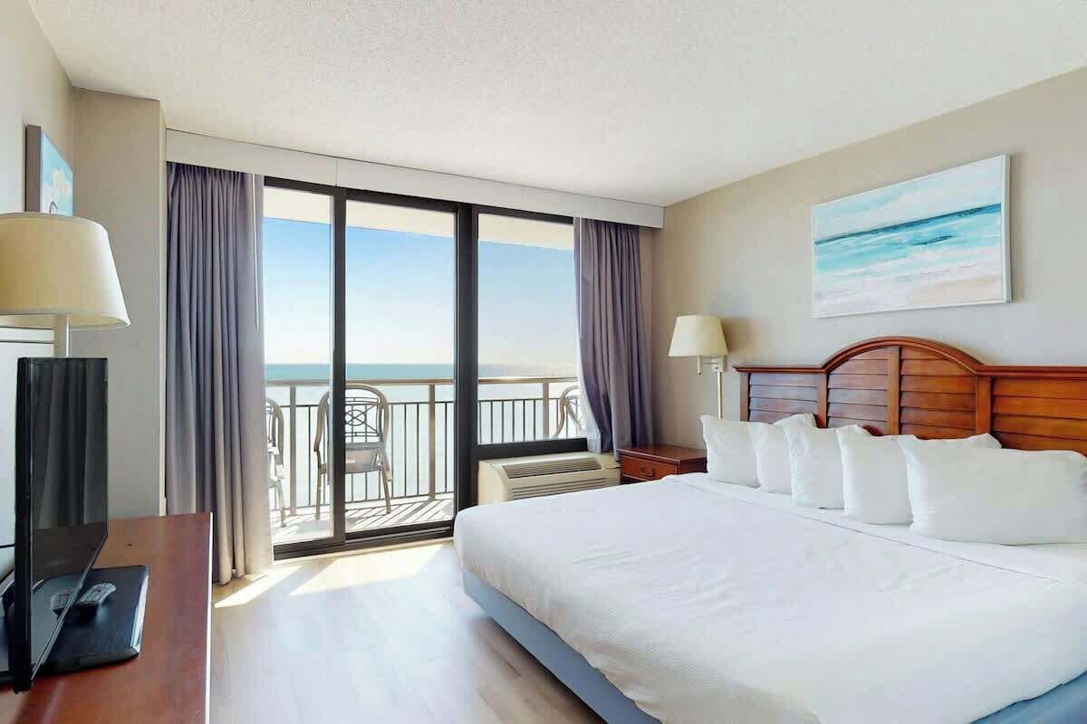 A hotel room with a large bed, a TV, a desk, and a balcony with a view of the sea.