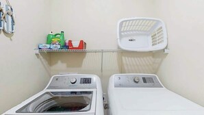 A laundry area with two washing machines, a shelf with laundry detergent, and a white laundry basket. - Corals and Oars Retreat (Myrtle Beach)