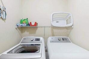 A laundry area with two washing machines, a shelf with laundry detergent, and a white laundry basket. - Corals and Oars Retreat (Myrtle Beach)