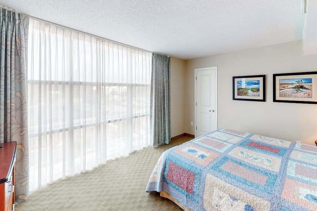 A bedroom with a large window, a bed with a quilt, a door, and two framed pictures on the wall.