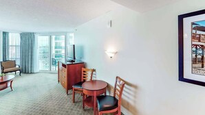 Quarto de hotel com área de jantar, televisão, quadro na parede e varanda com cortinas.