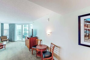 Ein Hotelzimmer mit Essbereich, einem Fernseher, einem gerahmten Bild an der Wand und einem Balkon mit Vorhängen.